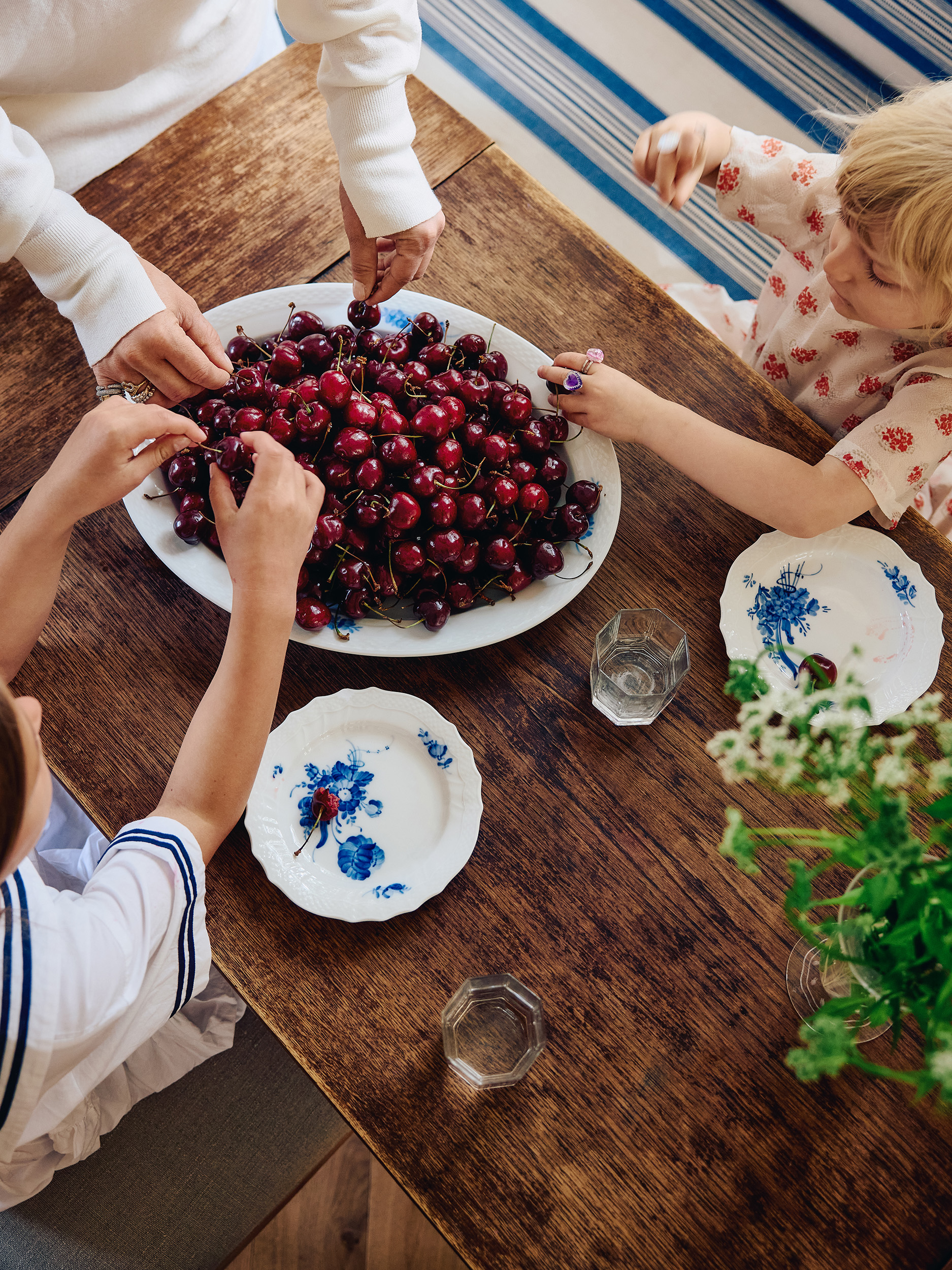 Top-down view of three people at a wooden table gathering fresh cherries from a large white serving platter.