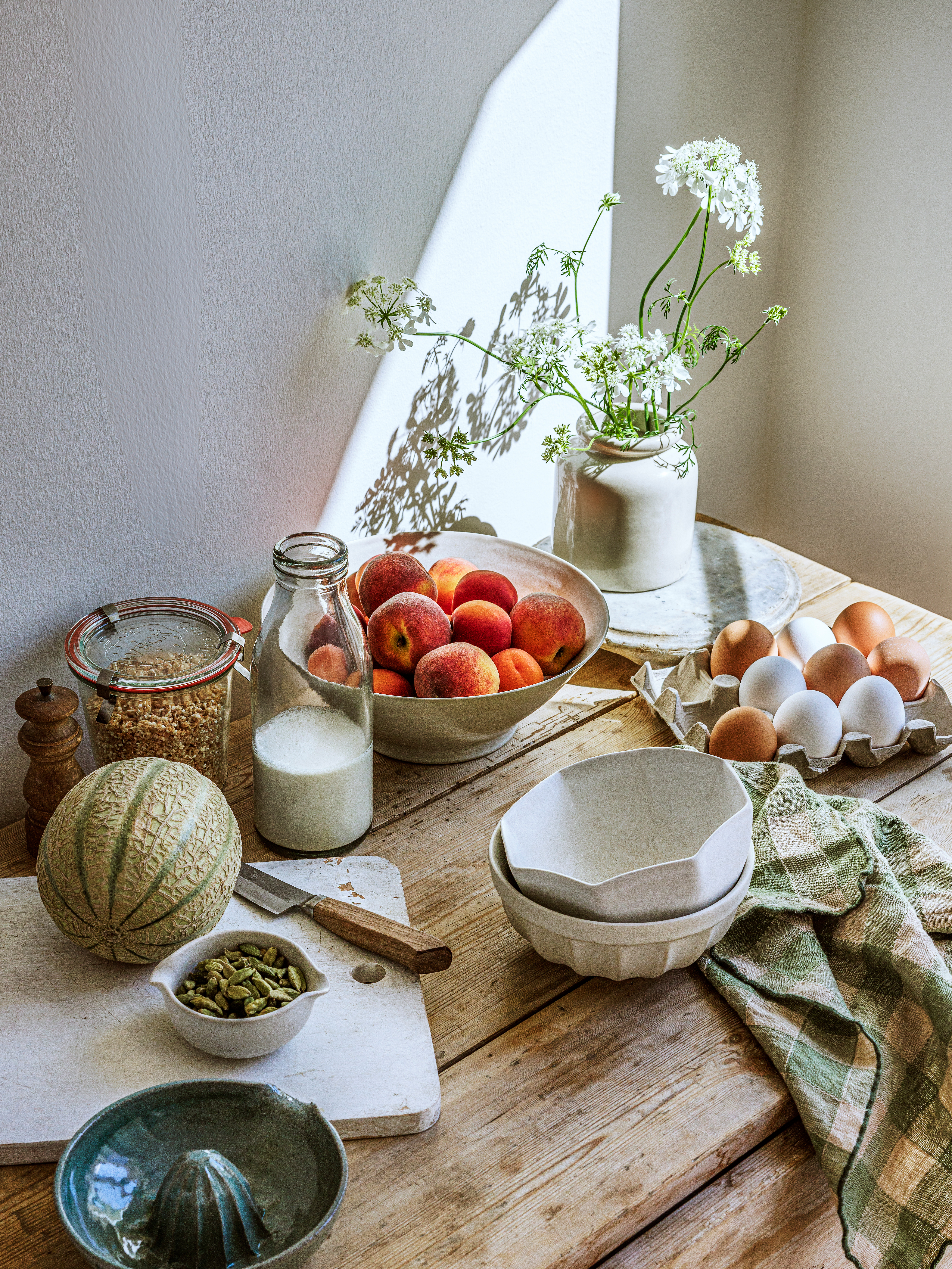 Still life on a wooden table with a bowl of peaches, milk bottle, melon, eggs, white flowers in a vase, and a green checked napkin.