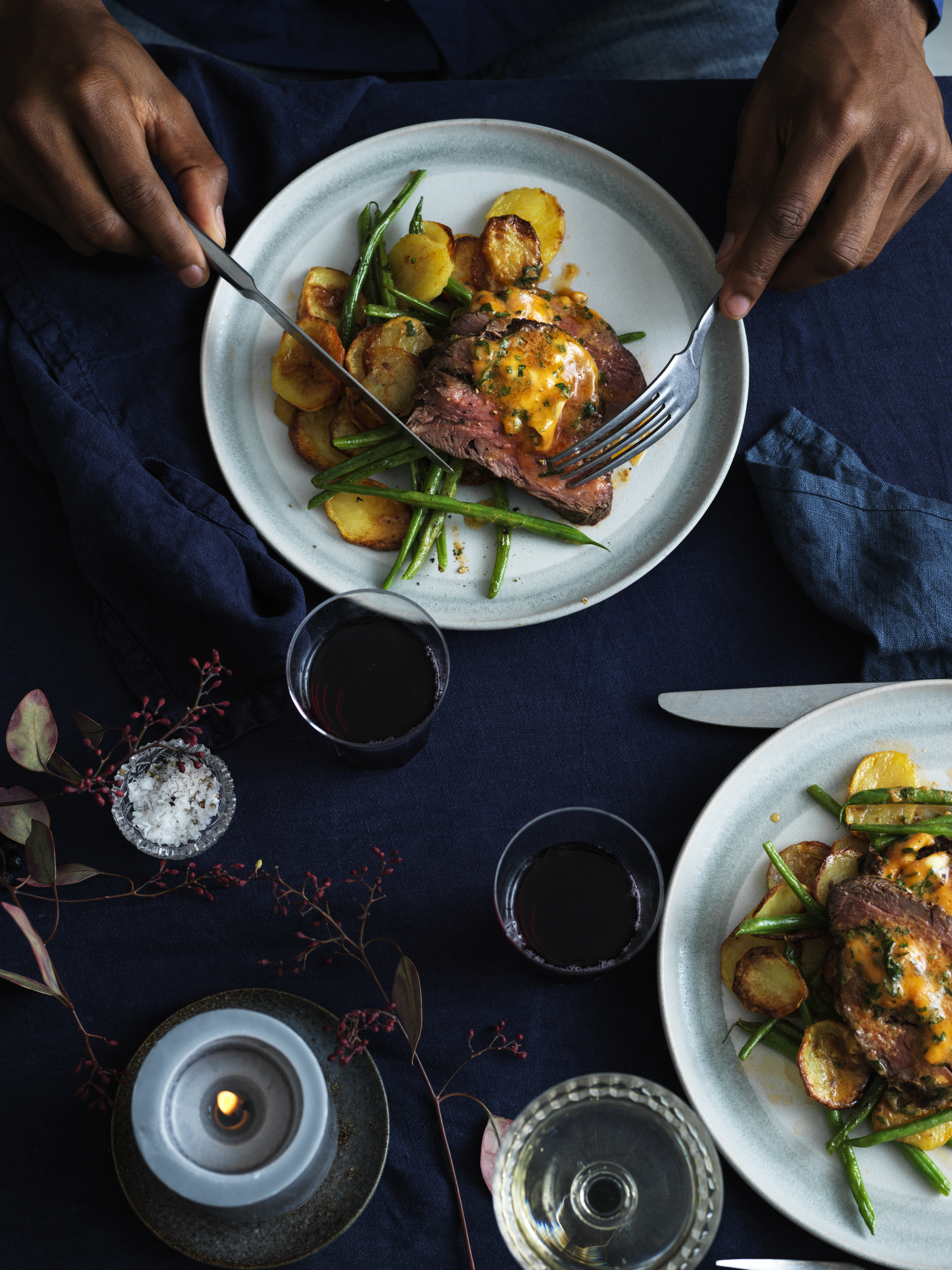 Person cutting a steak and green beans meal on a blue tablecloth with a burning candle and glass of red wine nearby