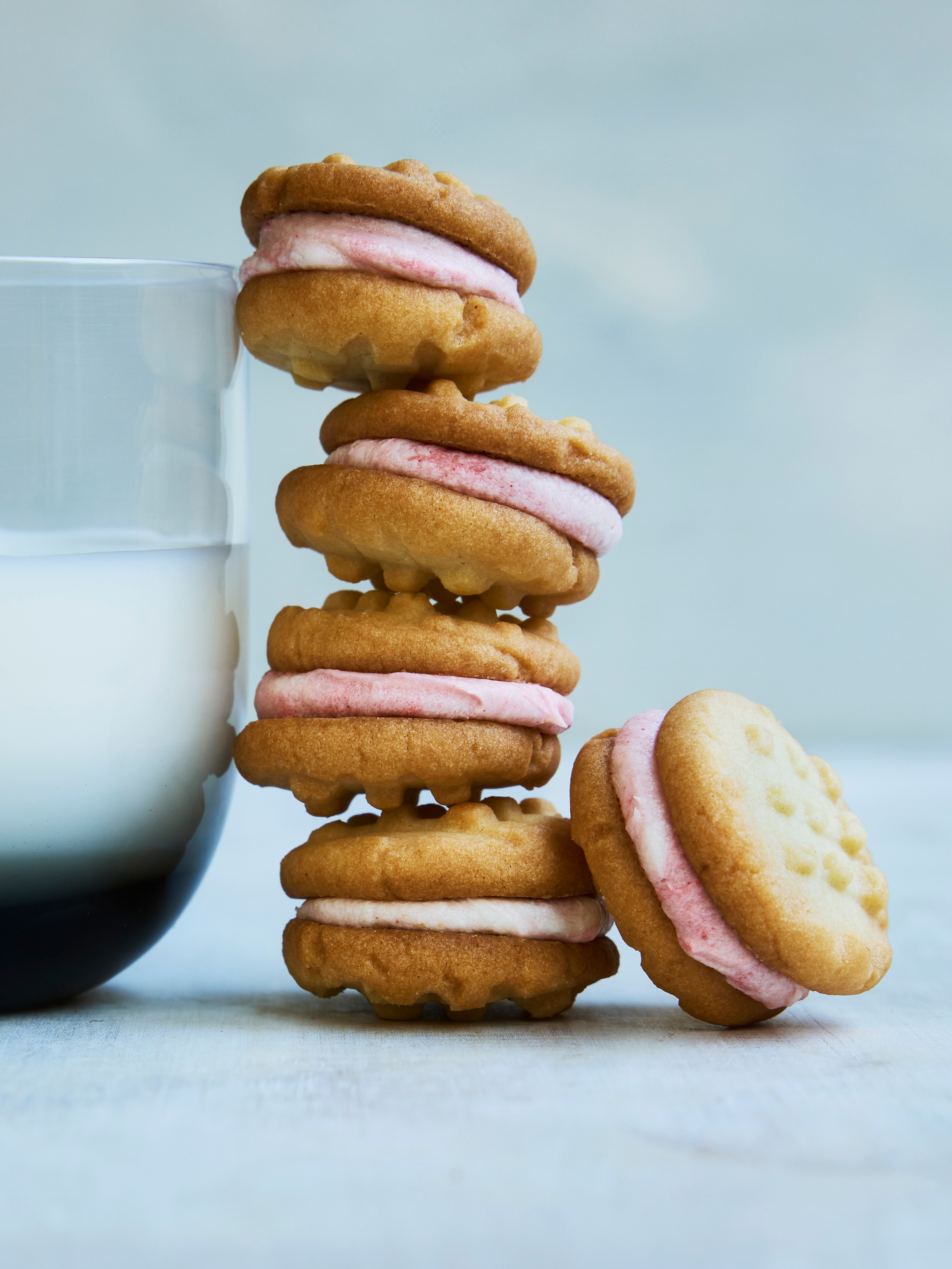 A stack of four round sandwich cookies with light pink cream filling beside a glass of milk on a light gray surface.