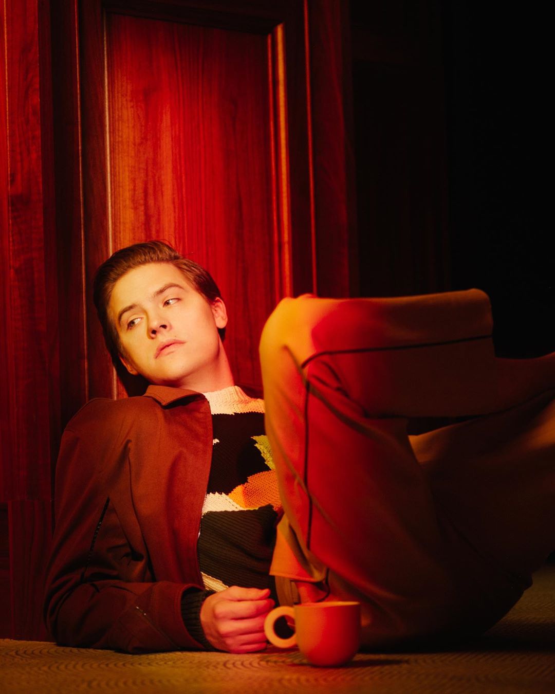 Portrait of an adult male lying on the floor in front of a red door with a mug in the foreground and wearing a brown jacket.