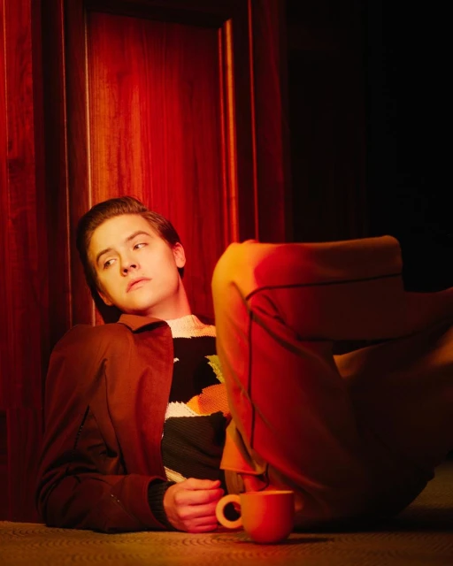 Portrait of an adult male lying on the floor in front of a red door with a mug in the foreground and wearing a brown jacket.