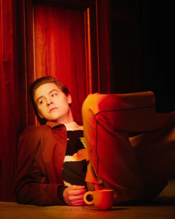Portrait of an adult male lying on the floor in front of a red door with a mug in the foreground and wearing a brown jacket.