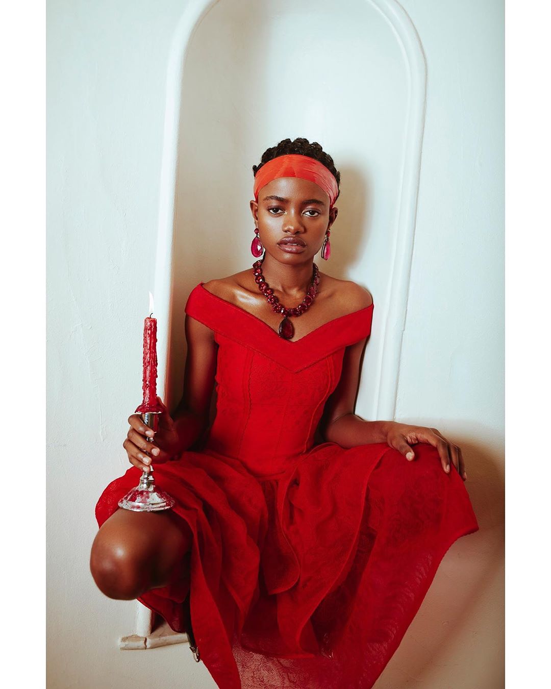 Elegant woman in a red dress poses in a bright indoor setting with a vintage bathtub, wearing statement jewelry and holding a lit red candle.
