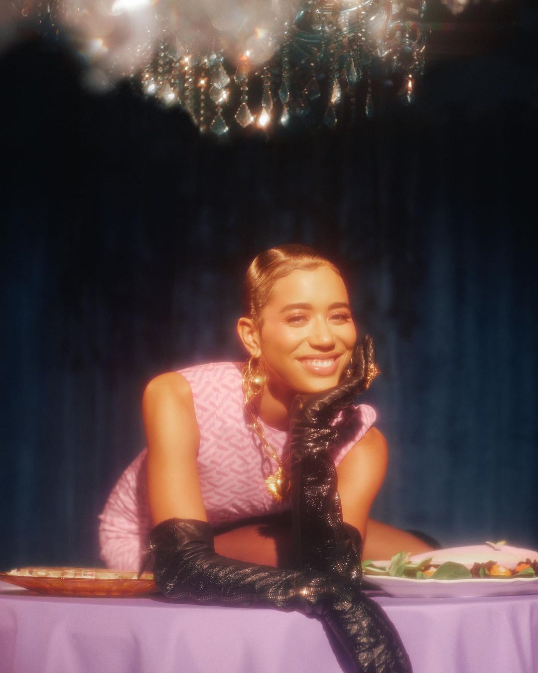 Glamorous woman at a lavishly decorated table, wearing vintage-inspired pink dress and elbow-length black gloves, smiling in a dimly lit space.