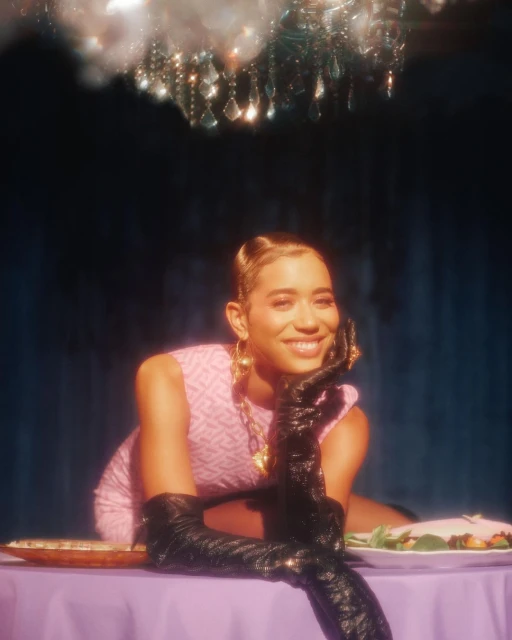Glamorous woman at a lavishly decorated table, wearing vintage-inspired pink dress and elbow-length black gloves, smiling in a dimly lit space.