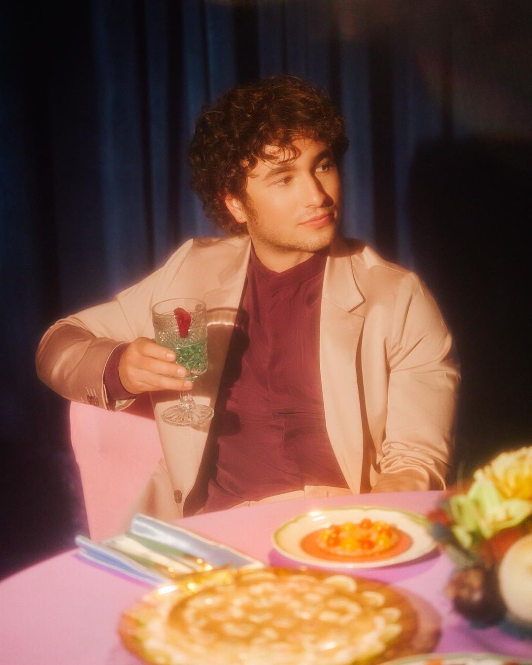 Man in a tan suit poses at a table with decorative food, drinks, and flowers, bathed in warm light against a blue curtain backdrop.