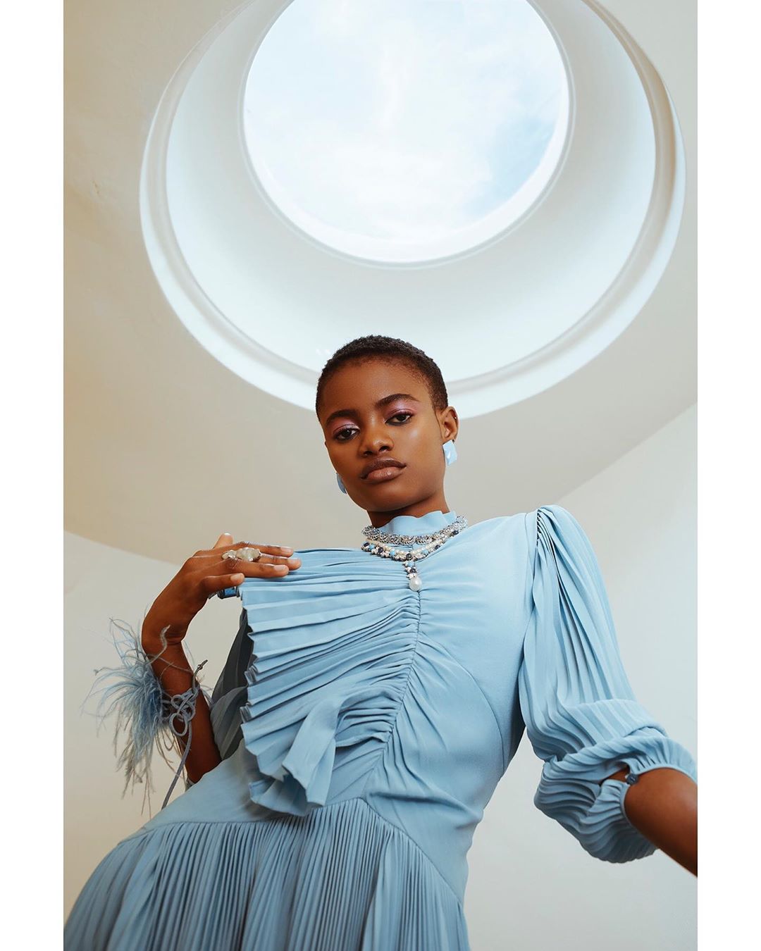 A young adult woman in a light blue dress with pleats and feather details poses in front of a circular skylight, looking down at the camera.