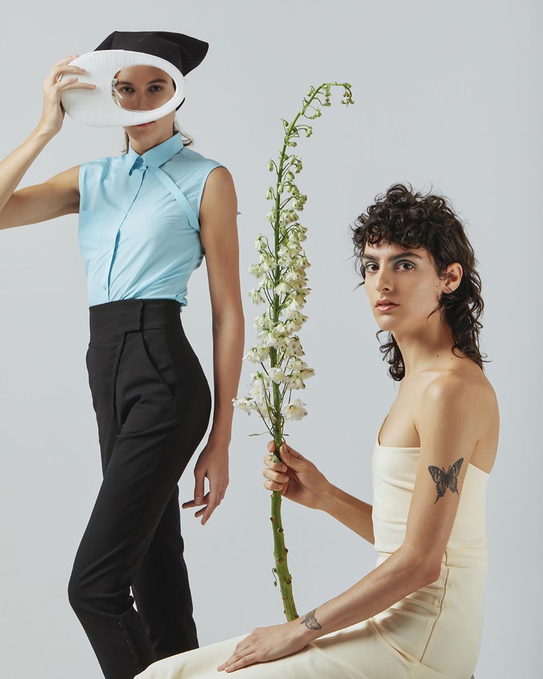 Two fashion models pose with a long white flower against a neutral backdrop. One woman wears a blue top, the other a cream dress.