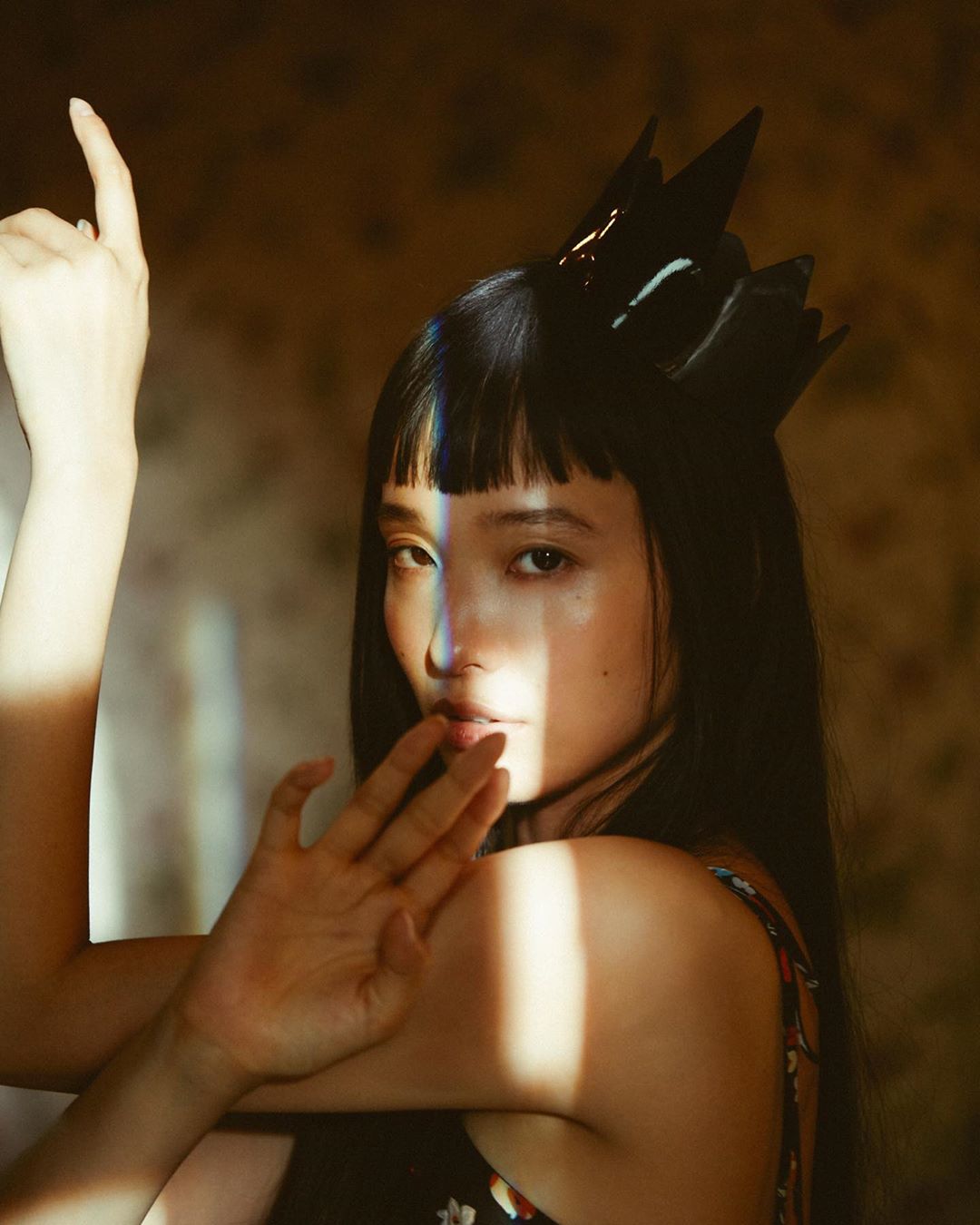 Portrait of an East Asian woman with black hair and a black crown, posing indoors with her hands up in a fashionable and artistic style.
