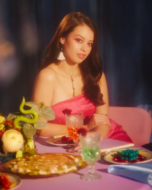A woman with dark hair sits at a table with food, cocktails and flowers in a staged indoor setting with pink and blue tones.