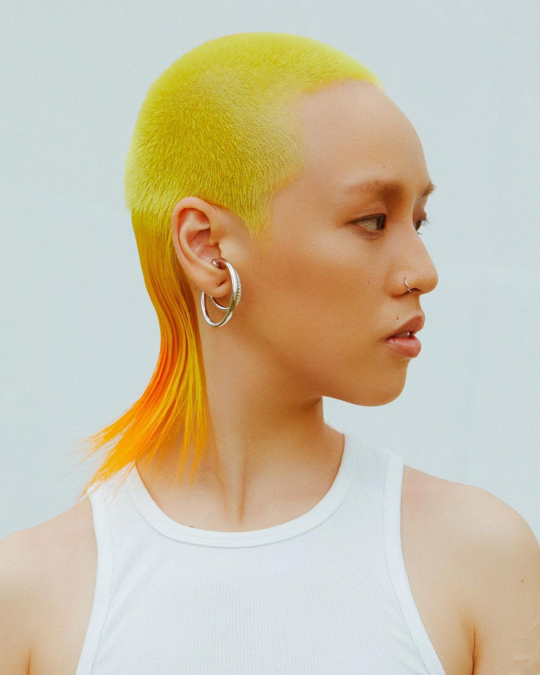 Portrait of a person with a yellow mullet haircut wearing a white tank top and hoop earrings against a pale blue background.