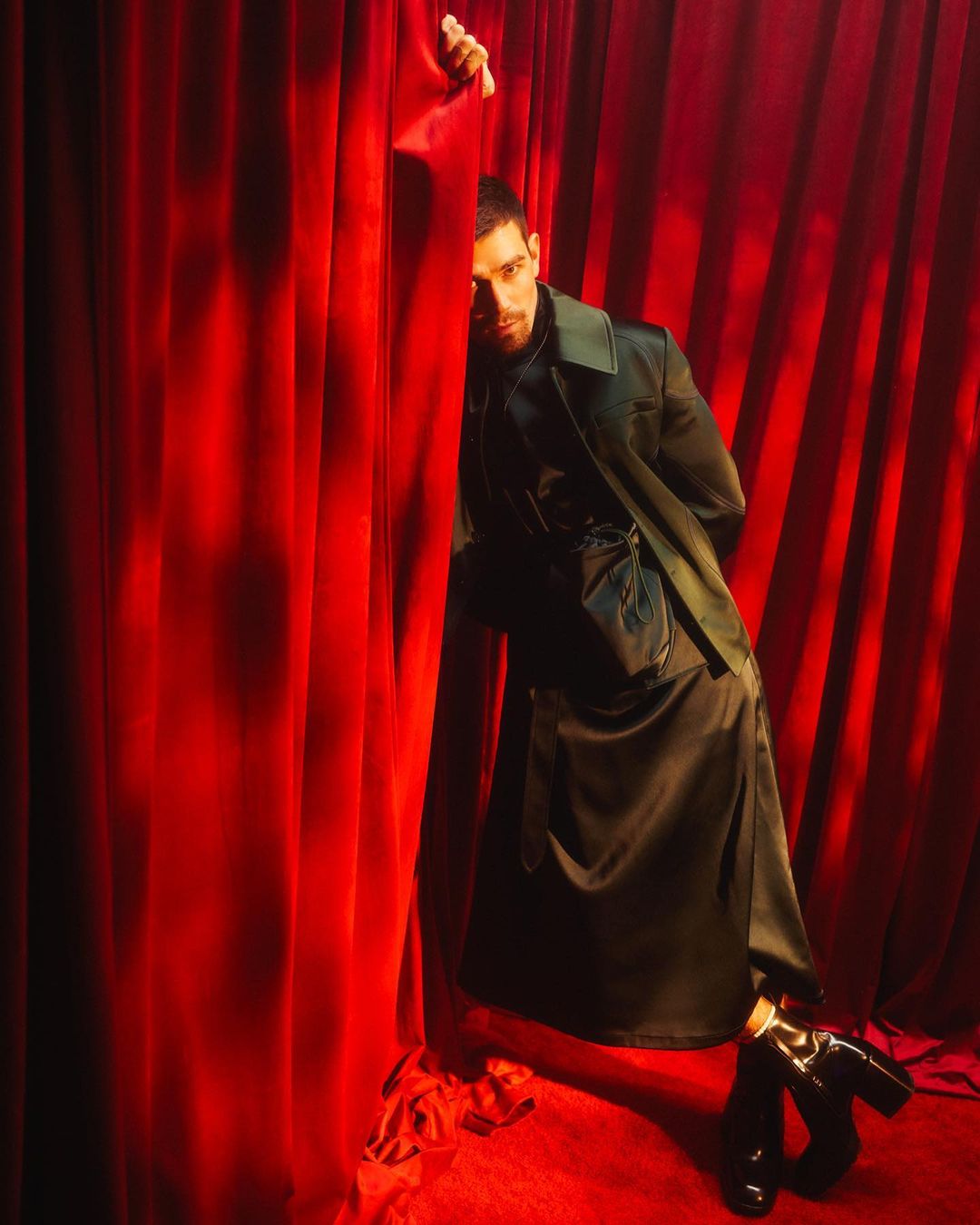 Portrait of an adult man standing by a red curtain. He is wearing a green jacket and black boots. Studio shot with dramatic lighting.