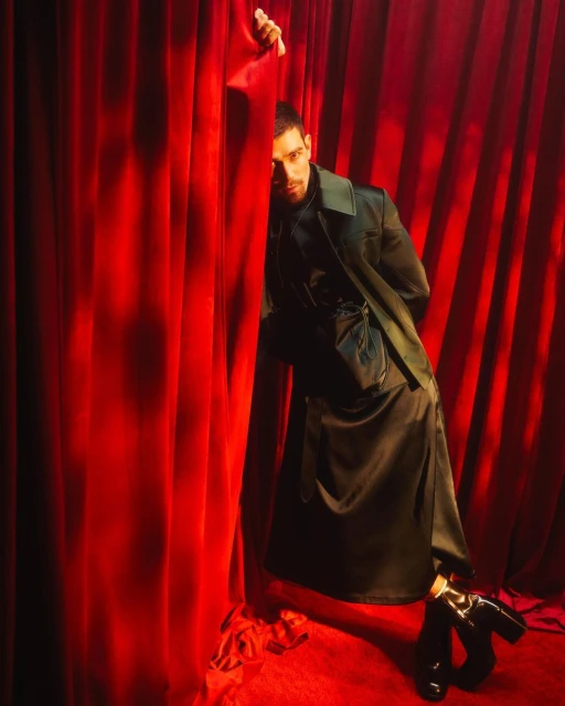 Portrait of an adult man standing by a red curtain. He is wearing a green jacket and black boots. Studio shot with dramatic lighting.