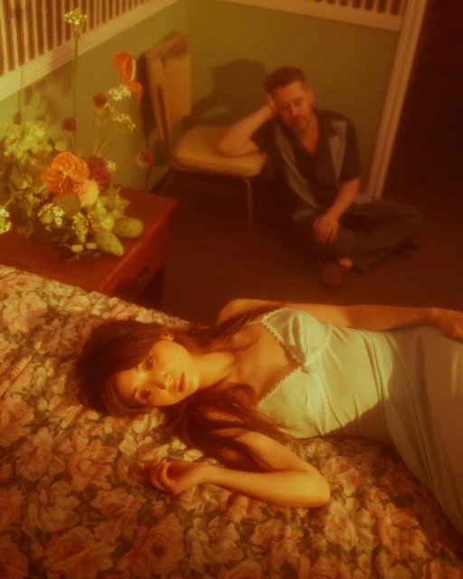 Woman in blue slip dress laying on floral bed with man seated in background. Moody lighting and warm color grading.