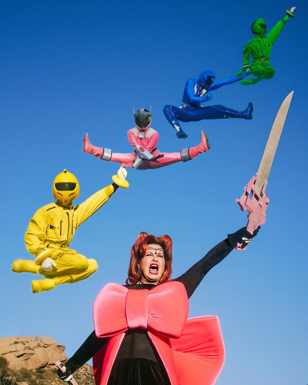 Group of costumed performers jumping against a blue sky with a large sword; mountains in the background; colorful outfits and dramatic poses.