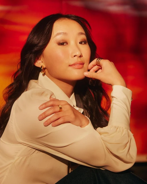 Portrait of an Asian woman with dark hair in a cream blouse with red and orange background. She is wearing rings and earrings.
