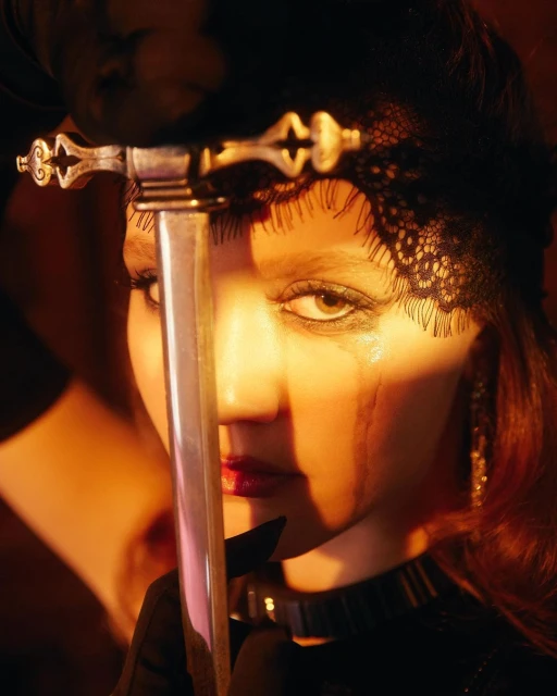 Dramatic close-up portrait of a woman with dark eye makeup, a lace headpiece, and a sword partially obscuring her face in warm-toned lighting.