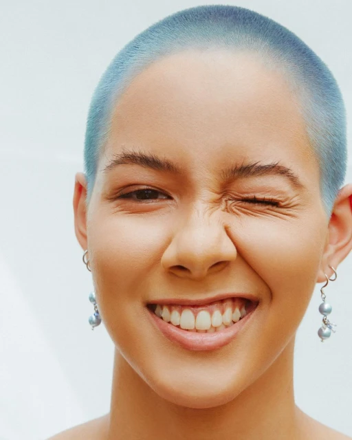 Close-up studio portrait of a young adult female with shaved, blue-tinted hair, smiling with visible teeth and winking, wearing silver earrings.