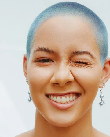 Close-up studio portrait of a young adult female with shaved, blue-tinted hair, smiling with visible teeth and winking, wearing silver earrings.