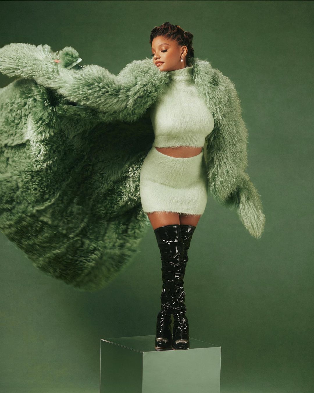 Fashionable woman in a fuzzy green two-piece outfit with a large fur coat and black patent leather thigh-high boots posing in a studio.