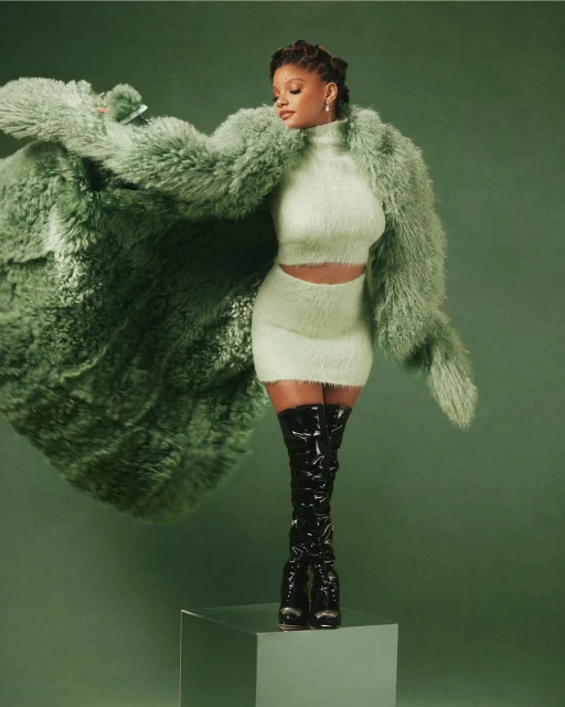 Fashionable woman in a fuzzy green two-piece outfit with a large fur coat and black patent leather thigh-high boots posing in a studio.
