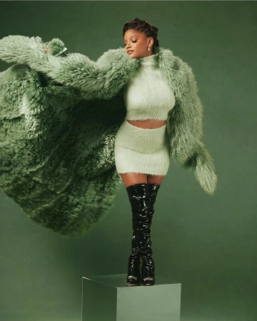 Fashionable woman in a fuzzy green two-piece outfit with a large fur coat and black patent leather thigh-high boots posing in a studio.