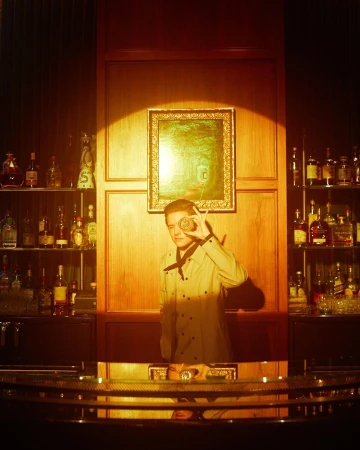 Bartender in dimly lit bar interior holding a cocktail glass to his eye, shelves of bottles and wood paneling in the background.