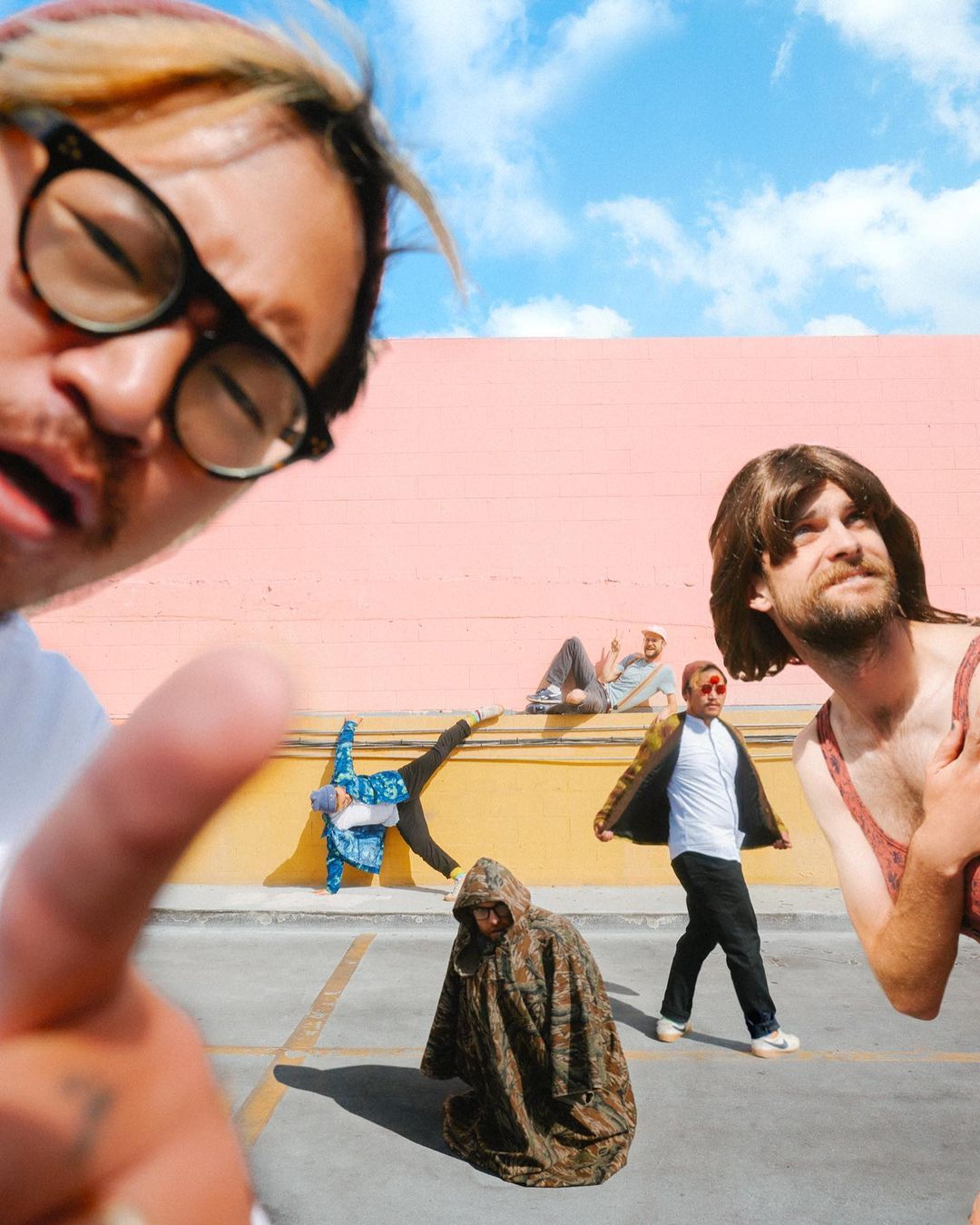 Group of people in an outdoor parking lot, with various poses and expressions, against a pink wall and blue sky.