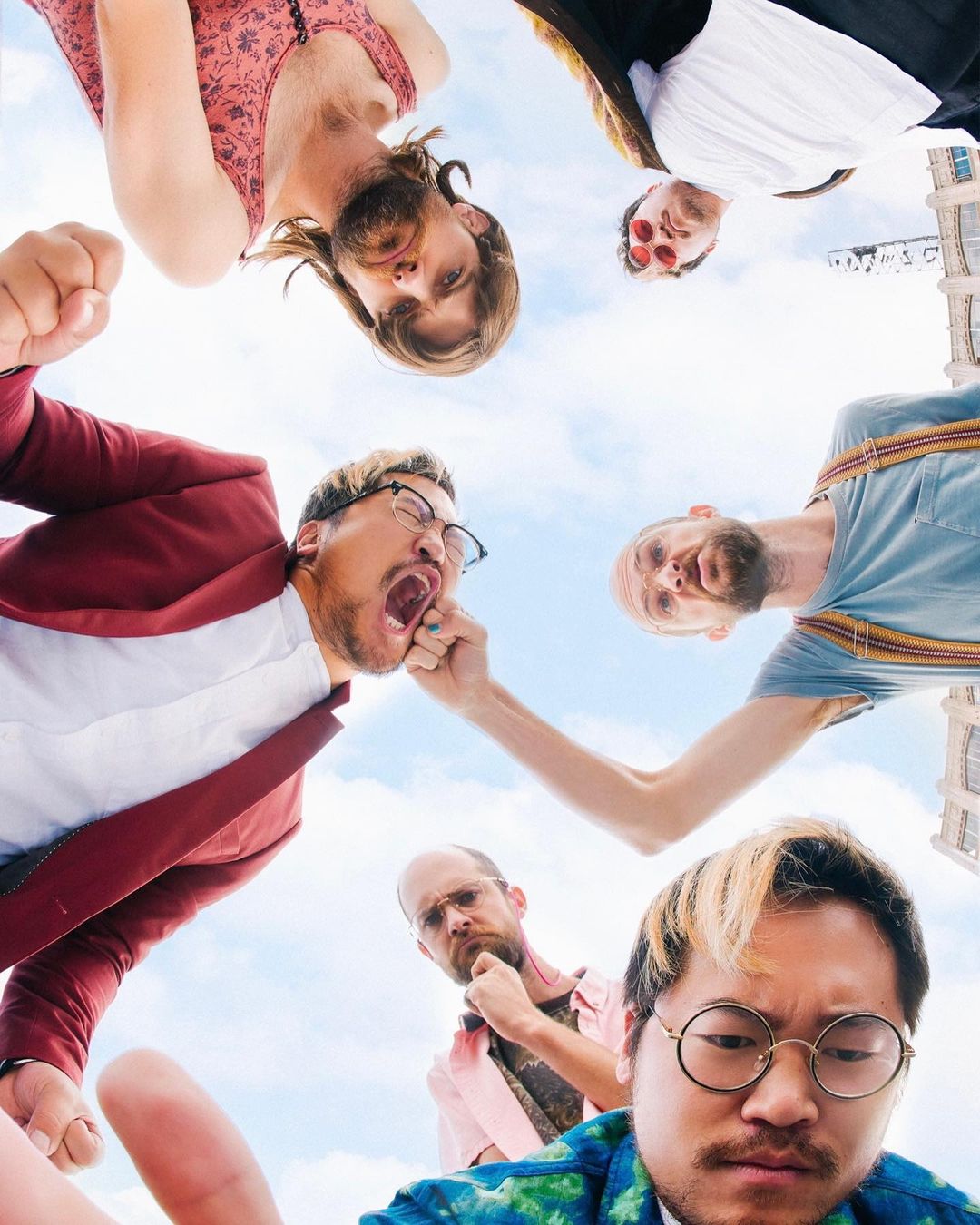 Group of six adults in casual clothing, looking down at the camera from an upward angle, with a blue sky and buildings in the background.