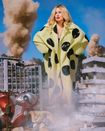 Adult female with blonde hair wearing a black and yellow polka dot coat in a destroyed, urban-like environment.