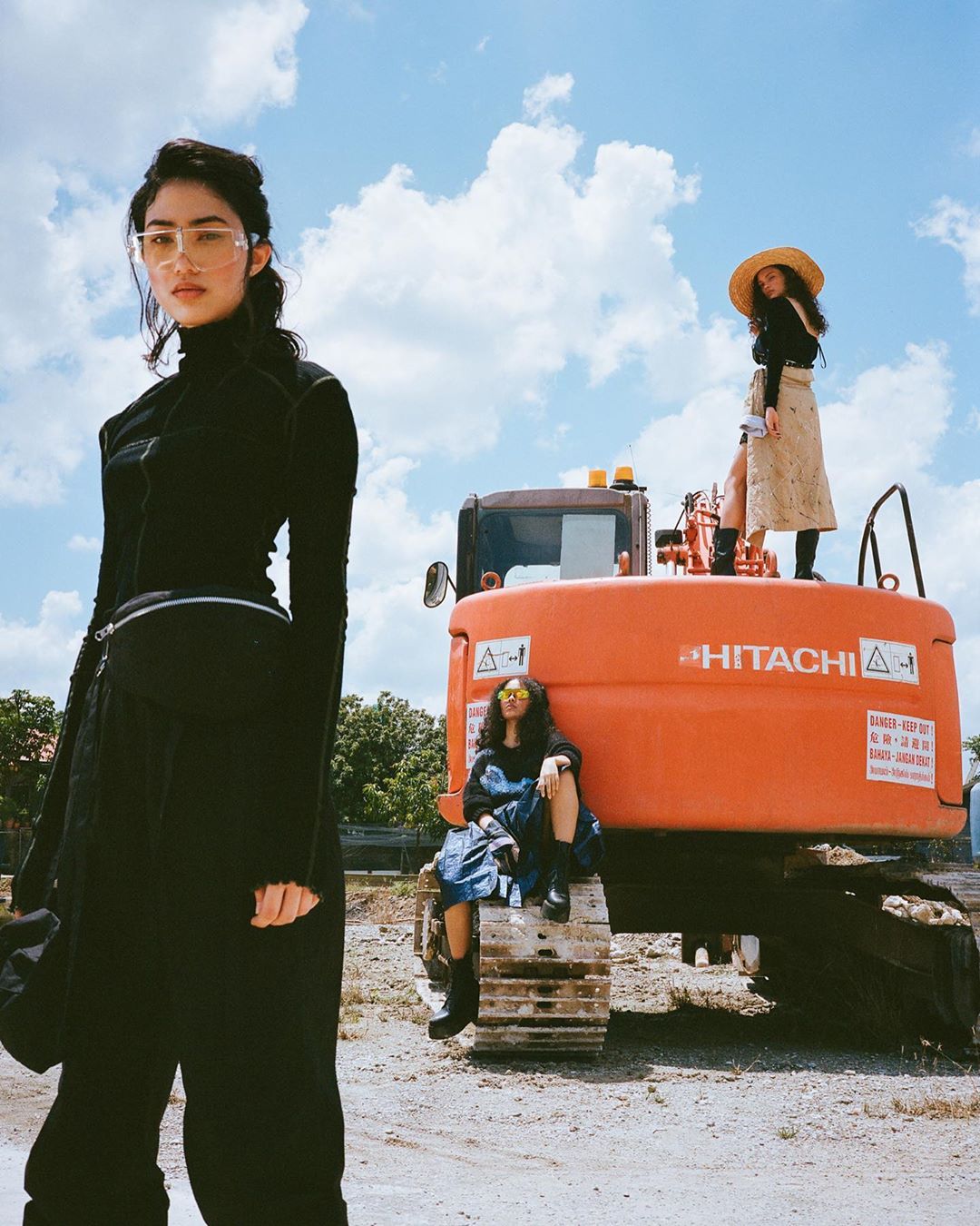 Three women pose on and around a large, orange construction tractor in a sunlit outdoor setting with a blue sky and clouds overhead.