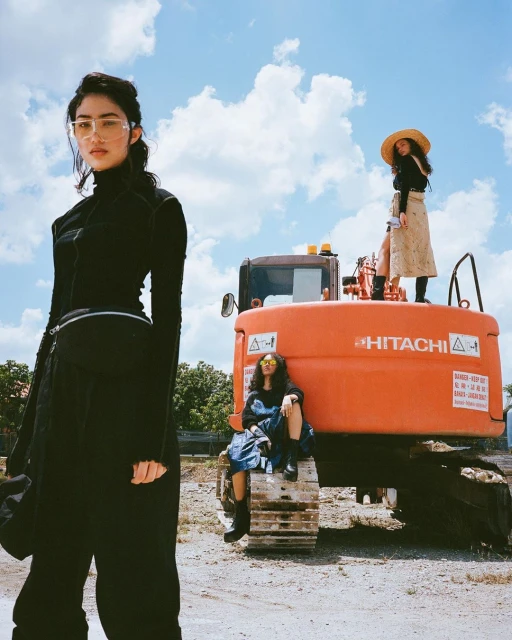Three women pose on and around a large, orange construction tractor in a sunlit outdoor setting with a blue sky and clouds overhead.