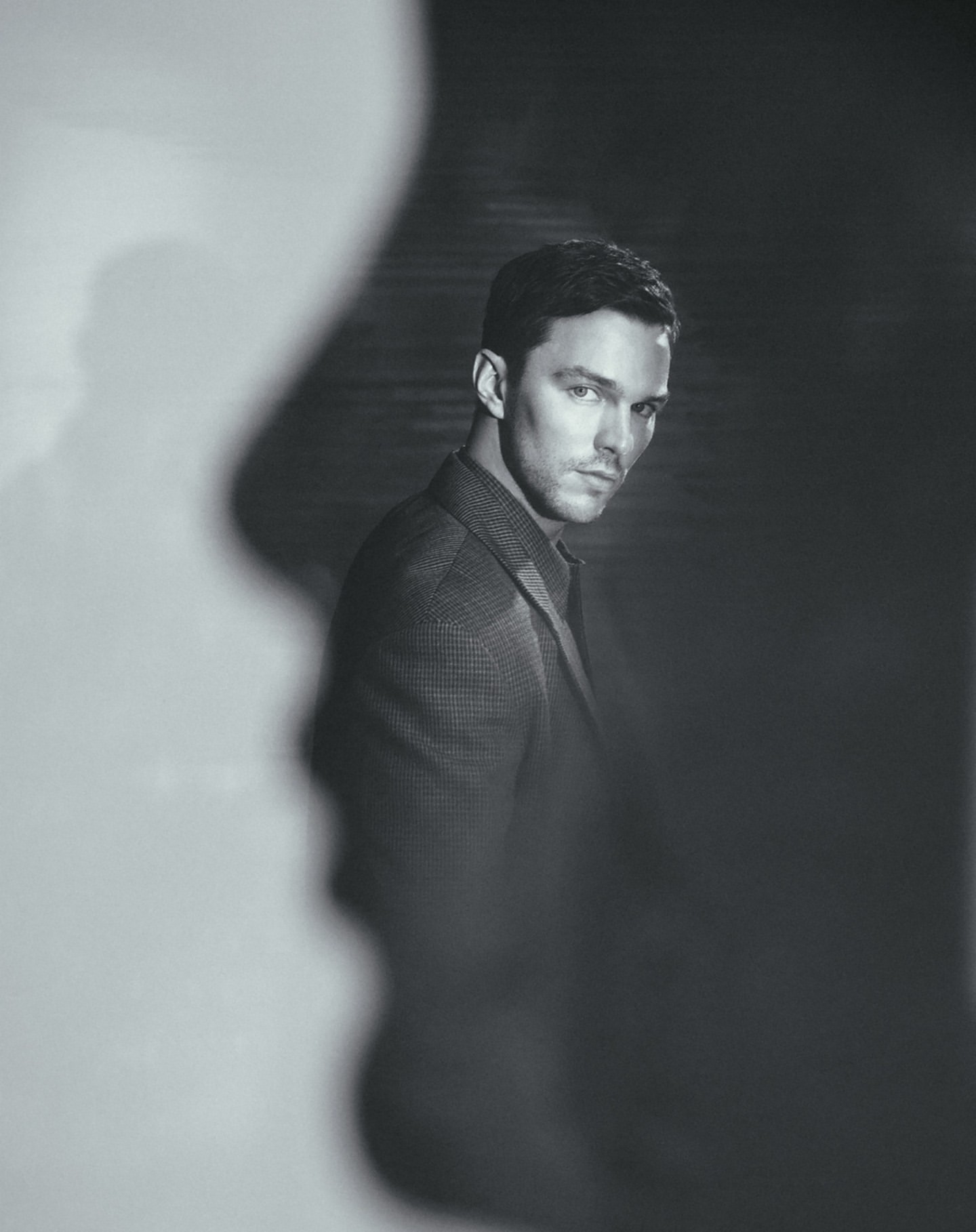 Black and white portrait of Nicholas Hoult wearing a suit jacket in a studio with light and shadow play for artistic effect.