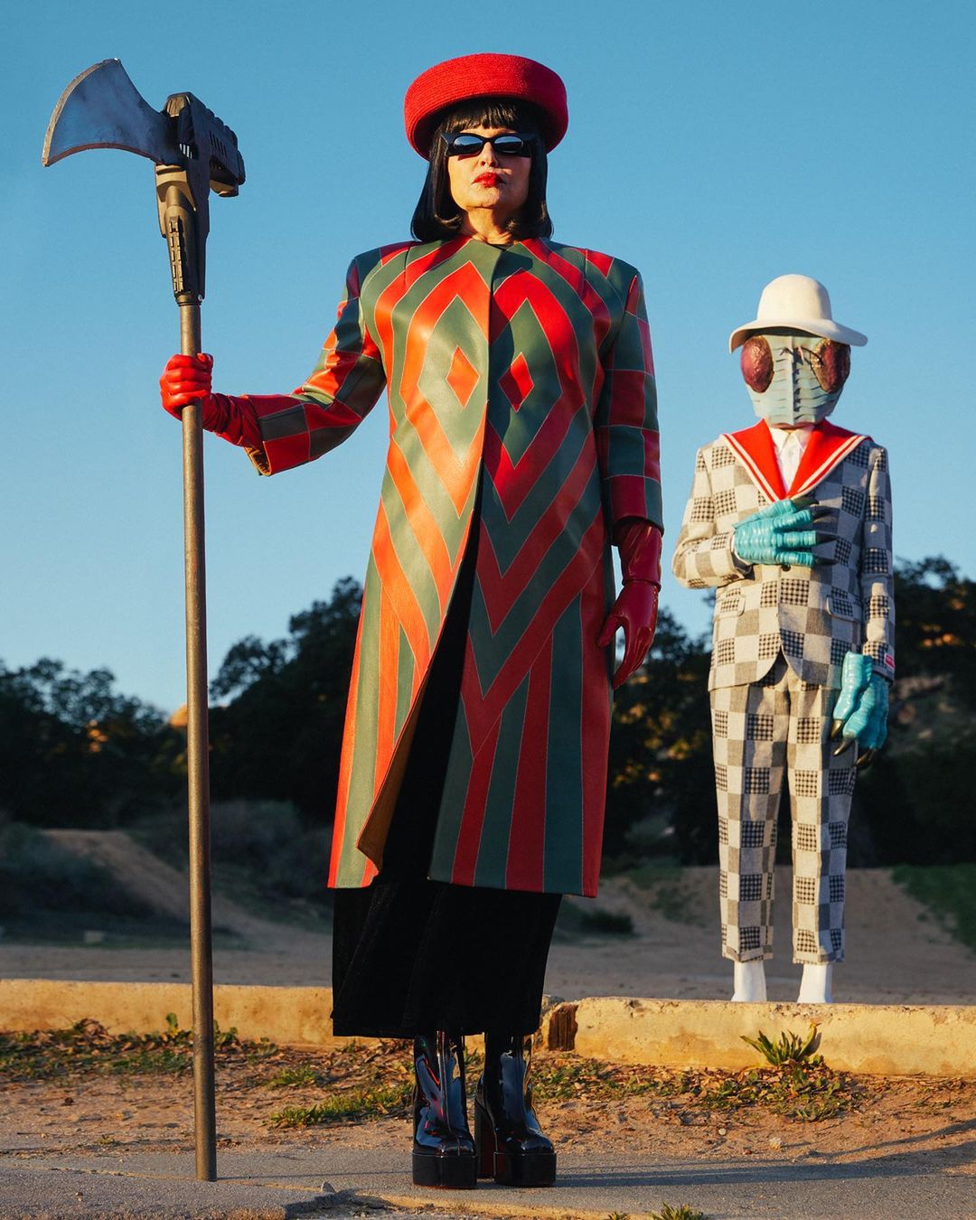 A woman in a geometrically patterned coat and sunglasses stands with an axe, next to a person in a fly-themed mask and suit, outdoors.