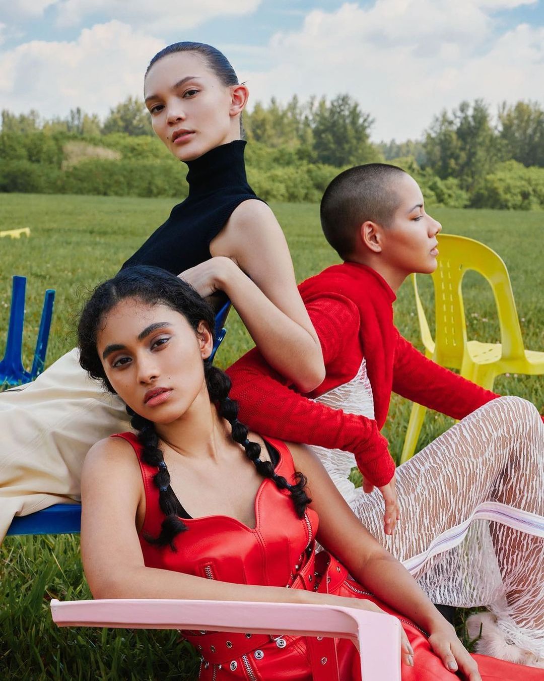 Three diverse women in red, white, and black high-fashion attire sit on colorful chairs outside in a grassy field beneath a blue sky.
