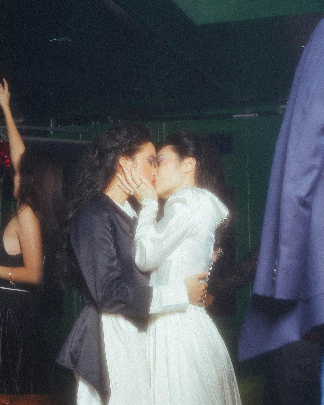 Two adult women embrace and kiss in a candid indoor shot. One is wearing a white dress, the other in a black jacket over a white dress.
