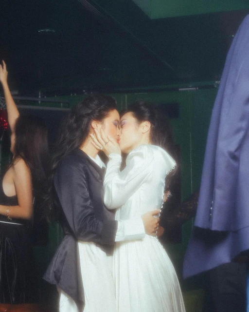 Two adult women embrace and kiss in a candid indoor shot. One is wearing a white dress, the other in a black jacket over a white dress.