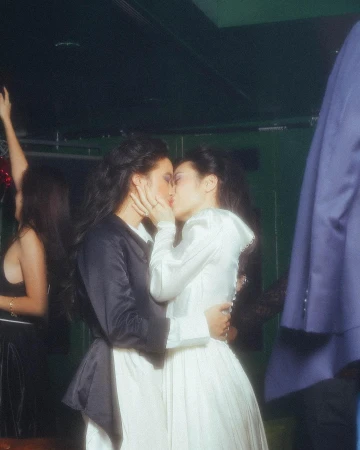 Two adult women embrace and kiss in a candid indoor shot. One is wearing a white dress, the other in a black jacket over a white dress.