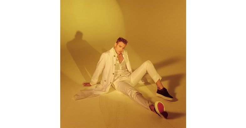 Blond male fashion model in white shirt, jacket, and pants, seated on warm yellow background with black-and-red sneakers.