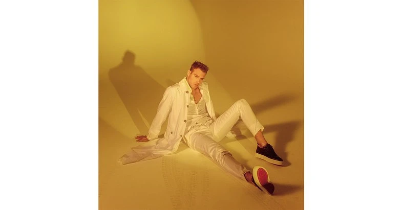 Blond male fashion model in white shirt, jacket, and pants, seated on warm yellow background with black-and-red sneakers.