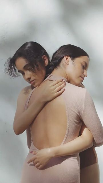 Two women in light pink dresses embracing in a studio with soft lighting and shadows creating an intimate and serene scene.