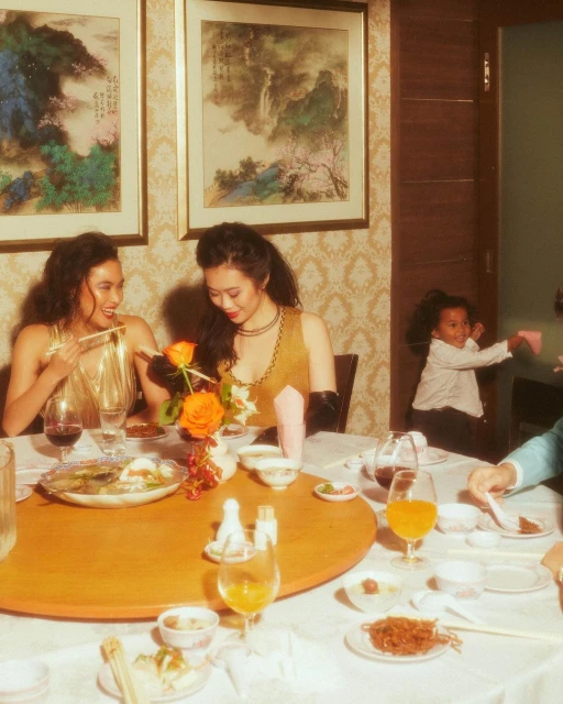 Two elegant women at a round table in a restaurant setting, enjoying food and drinks with a child in the background. Warm lighting.