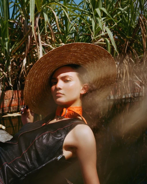 A woman wearing a straw hat and a black dress with orange collar poses in front of sugarcane plants in outdoor lighting.