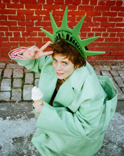 Young adult poses in a Statue of Liberty crown and light green trench coat, holding ice cream against a red brick wall backdrop.