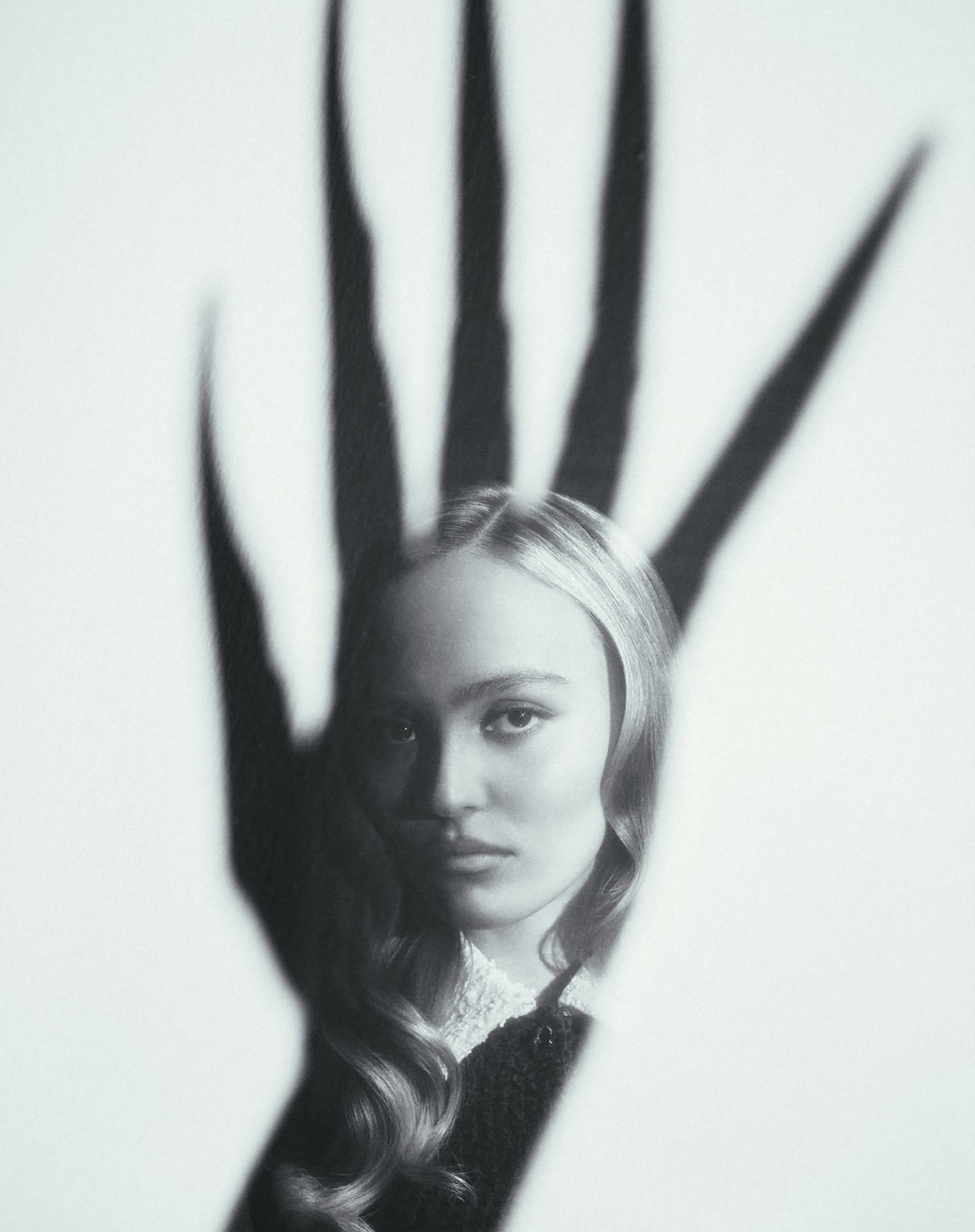 Monochrome portrait of a young woman with long wavy hair and a shadow of a hand with long fingers over her face on white.