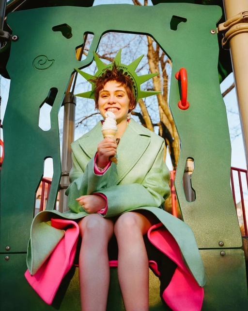 Young woman in Statue of Liberty crown and coat eating an ice cream cone seated in a playground, set outdoors in daylight.