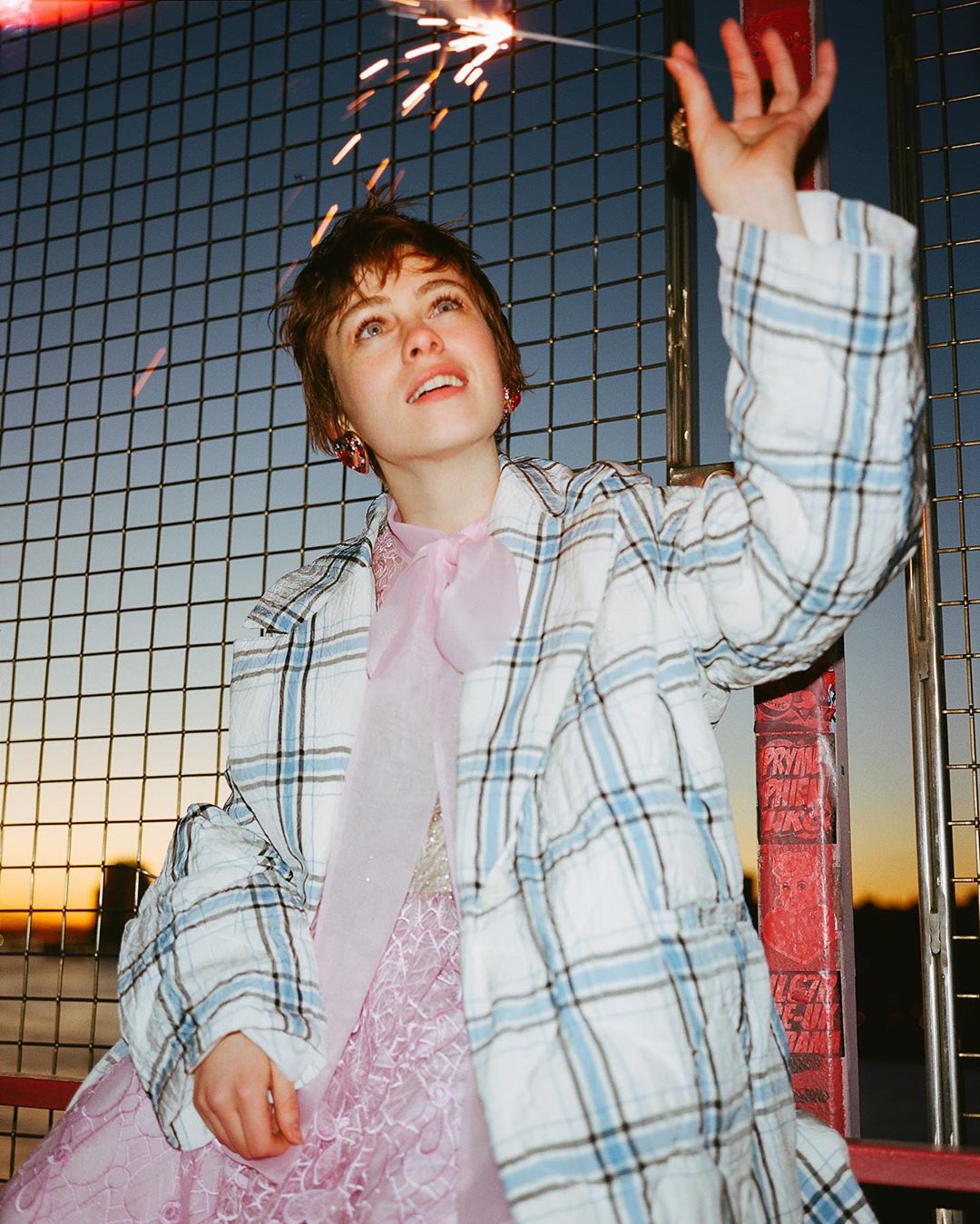 Woman in dress and jacket holds sparkler against blue sky and wire fence at dusk. Celebration imagery with vibrant colors.