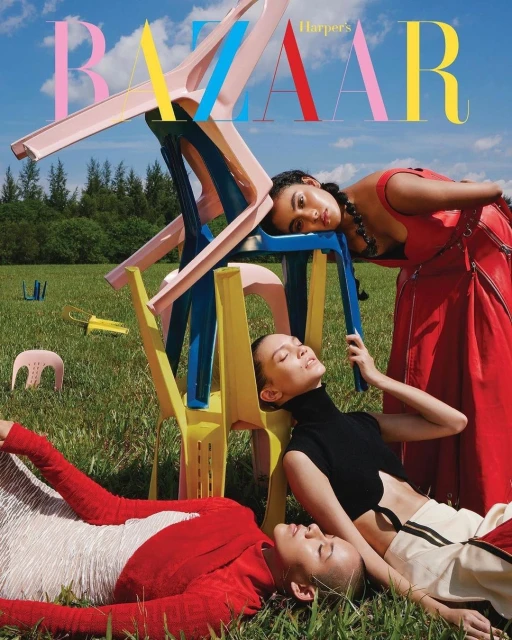 Fashion editorial cover featuring three women lying in grass, surrounded by stacked colorful chairs against sky and tree background. 