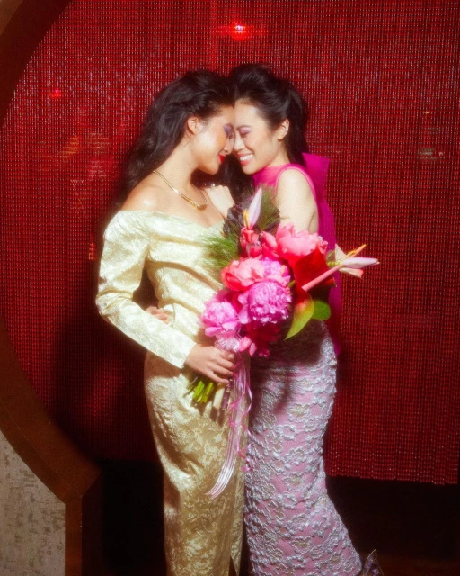Two women in gowns embrace near a red beaded wall, holding a pink peony bouquet, smiles, and formal wear for an elegant event.